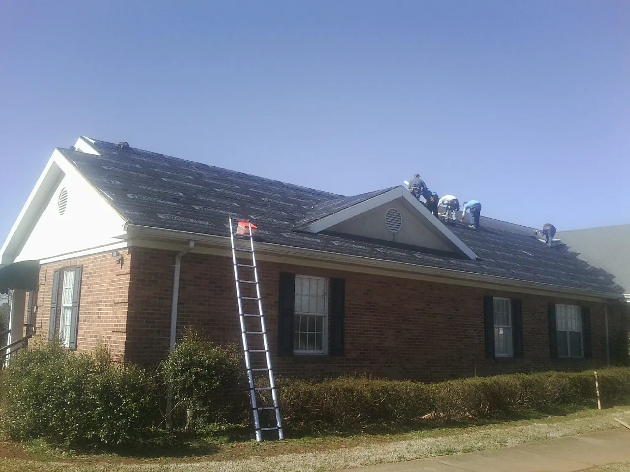 Crew tearing shingles off a brick ranch roof with a ladder set — Emergency Roof Repair work by our crew in Vona