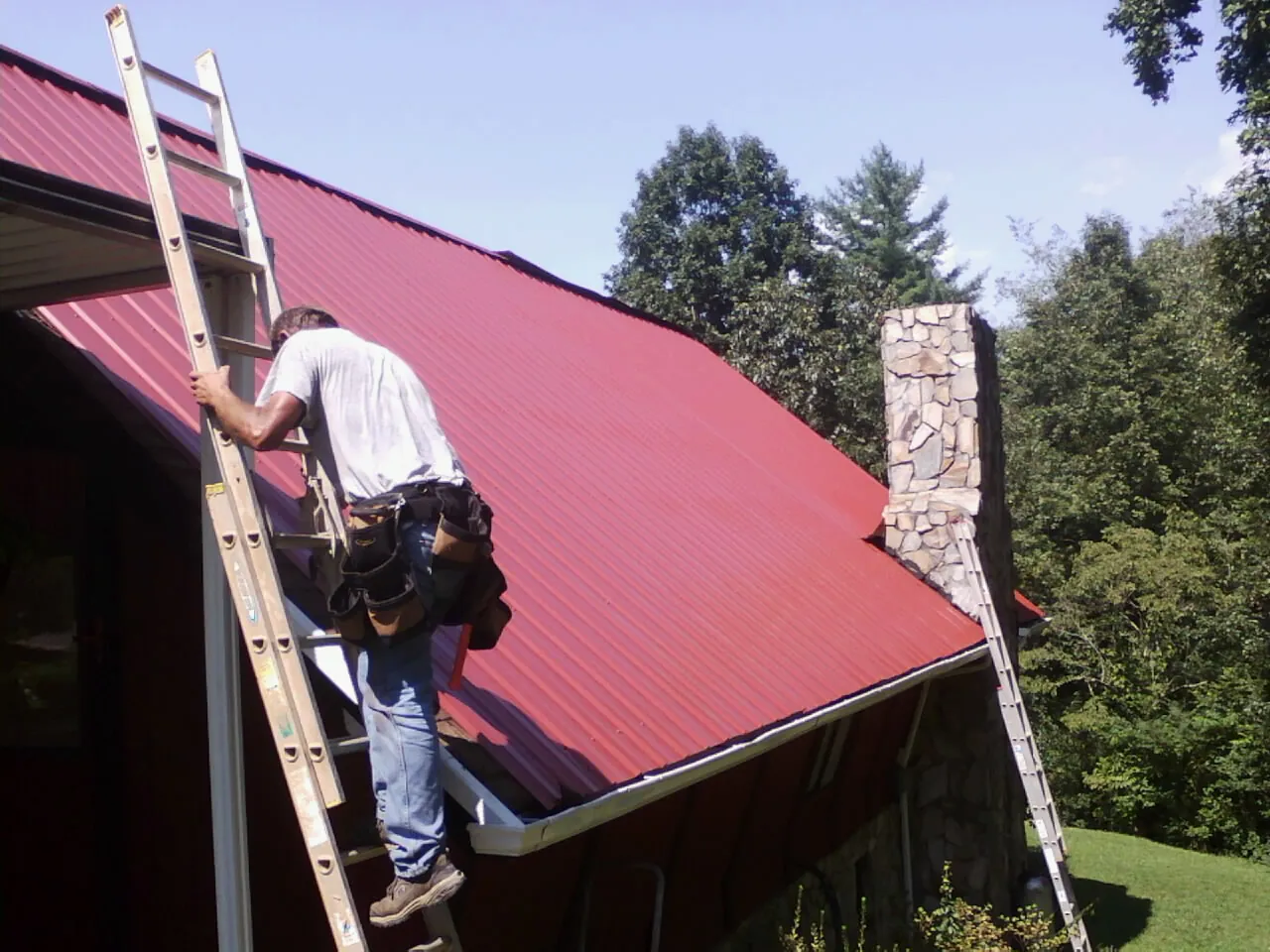 Roofer on a ladder beside a red metal roof and stone chimney — Soffit & Fascia Repair by Roofing Brothers in Vona