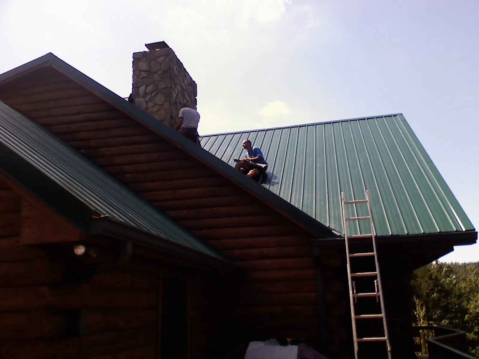 Roofers installing green metal panels beside a stone chimney on a log home in Vona